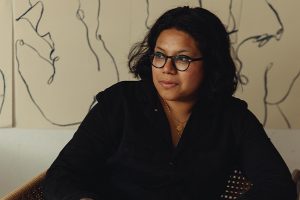 Artist Laxmi Hussain wearing black glasses and a black shirt sitting and looking away from the camera with a lined piece of art behind her on cream coloured paper