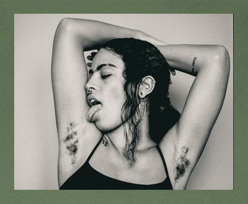 A black and while portrait of Aaliyah Ramsey with her arms above her head, proudly showing her underarm hair, with her head turned to the side, her eyes closed and her tounge out