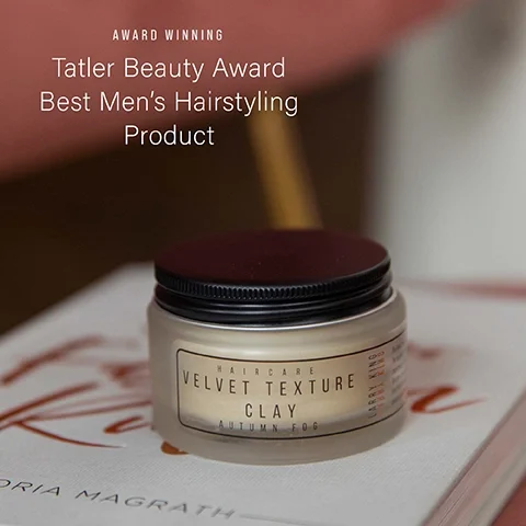 A jar of hair styling product sits on a book with overlaid text AWARD WINNING Tatler Beauty Award Best Mens Hairstyling Product and text on the jar label HAIRCARE VELVET TEXTURE CLAY AUTUMN FOG LARRY KING.
