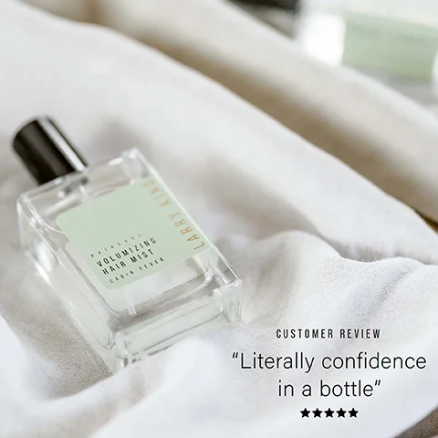 A clear spray bottle with a light green label reading Larry King Haircare Volumizing Hair Mist Cabin Fever lies on white fabric next to text Customer Review Literally confidence in a bottle and a five-star rating.