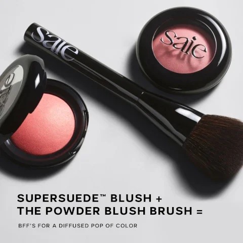 An overhead view of a round blush compact with a reddish shade, a brush with a black handle, and another round blush compact, featuring visible text that reads SUPERSUEDE™ BLUSH + THE POWDER BLUSH BRUSH = BFFS FOR A DIFFUSED POP OF COLOR.