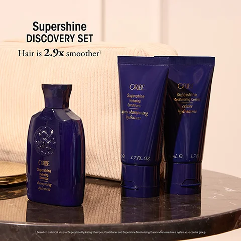 Three dark blue Oribe Supershine products including shampoo, conditioner, and moisturizing cream are displayed in front of a pouch with text that reads Supershine DISCOVERY SET Hair is 2.9x smoother.
