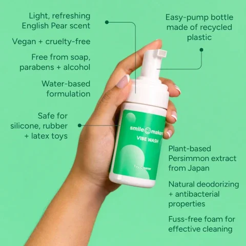 A person holds a white bottle labeled smile makers VIBE WASH with a green background, featuring text about the products features, including its scent, vegan formulation, and that it is safe for toy materials.