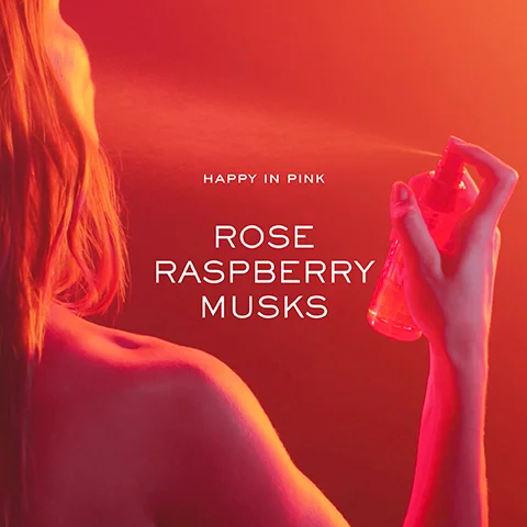 A woman with long hair is seen from behind, holding a pink spray bottle against a vibrant pink and orange background, with the text happy in pink, rose, raspberry, musks overlaid in white.