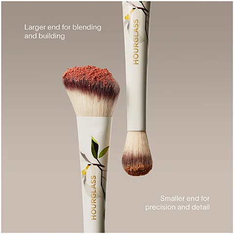 Two makeup brushes with white handles adorned with green leaves and yellow flowers, one with a large angled head and the other with a small rounded head, both covered in reddish-brown powder, are shown on a light background, with the text HOURGLASS, Larger end for blending and building, and Smaller end for precision and detail.