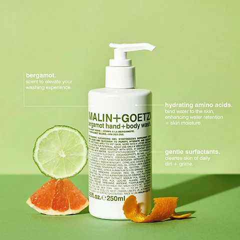 A white pump bottle of Malin+Goetz bergamot hand and body wash is displayed on a green background with a sliced lime, grapefruit, and orange peels, featuring visible text that reads MALIN+GOETZ bergamot hand+body wash 8.5 FL.OZ. 250 ML, bergamot. scent to elevate your washing experience., hydrating amino acids. bind water to the skin, enhancing water retention + skin moisture., gentle surfactants. cleanse skin of daily dirt + grime.